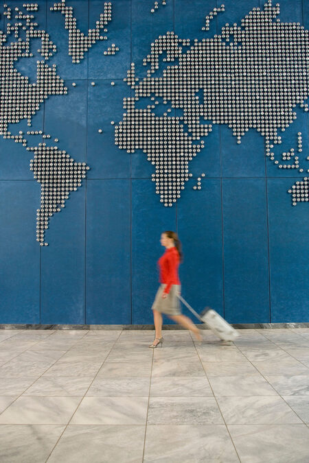 Woman walking past a wall with the world map on while pulling luggage behind her